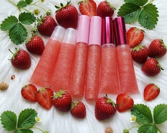 May include: Six clear squeeze tubes of pink glittery lip gloss arranged in a row. Each tube has a cap, with two light pink, two dark pink, and two clear caps. The tubes are surrounded by fresh red strawberries and green leaves.