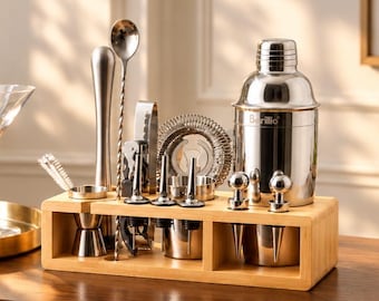 May include: A stainless steel cocktail shaker set with a wooden stand. The set includes a shaker with the text Barillio, a jigger, a long mixing spoon, a strainer, pourers, and a muddler. The tools are organised in a rectangular bamboo holder.
