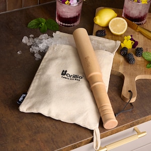 May include: A wooden ice mallet with a canvas bag labeled "Barillio Lewis Ice Bag". The bag is filled with ice cubes and the mallet is resting on top of the bag. There are lemons, blackberries, and flowers in the background.