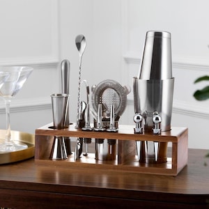 May include: A stainless steel bar tool set displayed in a wooden holder. The set includes a cocktail shaker, jiggers, a strainer, a bar spoon, and other tools. The tools are arranged on a dark brown wooden surface.