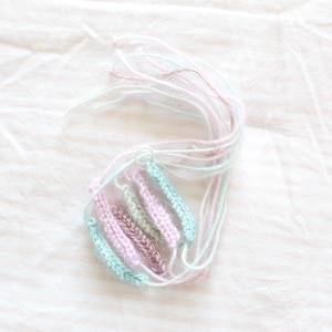 Puede incluir: Un conjunto de pequeños segmentos rectangulares de ganchillo en tonos pastel de azul claro, rosa suave y verde apagado. Cada segmento está unido a un largo y fino hilo de lana blanco, creando un grupo de delicados componentes de fibra texturizada.