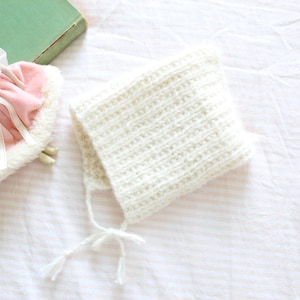 May include: A cream-colored, ribbed knit bonnet featuring two thin ties at the chin. Beside it sits a small doll wearing a pink dress with a white faux fur trim, a pink rose accent at the chest, and a sheer white veil attached to the head.