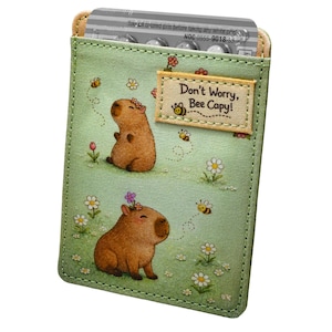 May include: A light green pill case with a capybara illustration and the text "Don't Worry, Bee Capy!". The case features a floral design with bees and flowers. The top of the case has a beige trim and contains a pill pack.