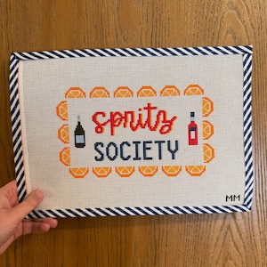 May include: A needlepoint canvas with the words "Spritz Society" in red and navy blue, surrounded by orange slices. The canvas has a navy and white striped border and includes illustrations of a bottle and a liquor bottle.