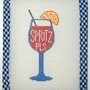 May include: Needlepoint art piece featuring a cocktail glass design. The glass is filled with red liquid and the words "SPRITZ PLS" in blue. An orange slice and a straw are included. The piece is framed with a blue and white gingham border.