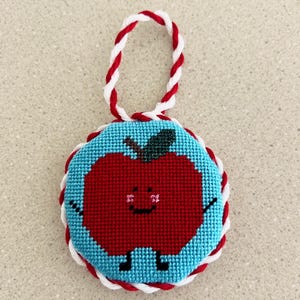 May include: A handmade, cross-stitched apple ornament. The red apple has a smiling face, black legs, and arms, set against a light blue background. The ornament is trimmed with red and white cording and has a twisted red and white cord for hanging.