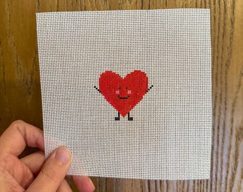 May include: A cross-stitch canvas featuring a smiling red heart with arms and legs. The heart is centered on a white, grid-patterned canvas. The canvas is held by a hand, with a wooden surface in the background.