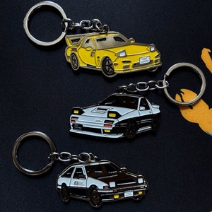 May include: Three car-shaped keychains. The top keychain is yellow, the middle one is white and black, and the bottom one is white and black. Each keychain has a silver keyring attached. The background is dark.