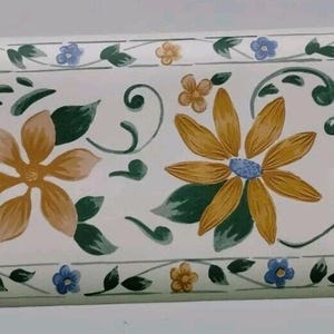 May include: Floral wallpaper border featuring a repeating pattern of stylized flowers and leaves. The design includes large blue and yellow flowers, green leaves, and smaller yellow and blue blossoms, all set against a white background.