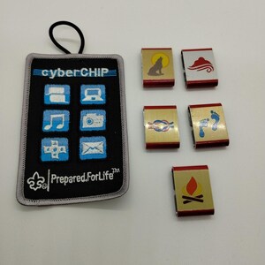 May include: A black rectangular embroidered patch with a black elastic loop, featuring the text cyberCHIP and Prepared.ForLife, alongside six blue icons. Five small rectangular metal slides with red edges and gold faces display symbols of a wolf, cloud, knot, footprints, and campfire.