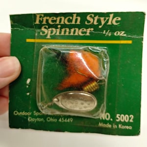 May include: A French Style Spinner fishing lure weighing 7 grams in its original green packaging. The lure features a bright orange fuzzy body, a gold-coloured ribbed bead, and a silver-toned metal blade. The packaging includes the text No. 5002.