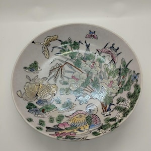 May include: A round, off-white ceramic plate with a detailed, colorful floral and bird design. The plate features a central image of a bird, fish, and flowers, with butterflies and dragonflies. The rim is a pale, neutral color.