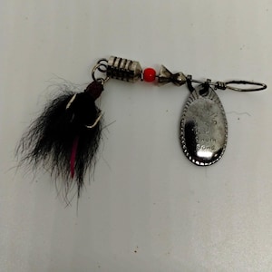 May include: A fishing lure featuring a silver-toned metal spinner blade engraved with the text South Bend. The lure includes a black feathered hook, a small red bead, a ribbed cylindrical metal weight, and a conical metal component on a wire.