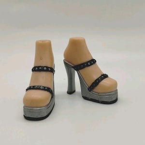 May include: A pair of doll feet wearing silver platform high-heeled sandals. The sandals feature two thin black straps across the top of the foot, each adorned with small silver studs. The platform soles are metallic silver with a black base.