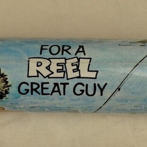 May include: A cylindrical gift wrap featuring a cartoon illustration with a fishing theme. The design includes a wolf character, a fishing rod, and the text "FOR A REEL GREAT GUY" in a playful font, set against a blue and green backdrop.