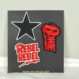May include: A set of three embroidered iron-on patches on a black backing card labeled IRON-ON PATCH SET. The patches include a black star with a white border, a red patch with the word Bowie, and a red patch with the text REBEL REBEL.