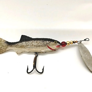May include: A fishing lure shaped like a fish with a black back, silver sides, and a textured tail. It features a red mouth, a yellow eye, a triple hook on the underside, and a metal spinner blade attached to the front with red and gold beads.