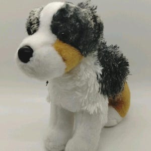 May include: A plush dog toy featuring a tri-colour coat of white, black, and tan fur. The stuffed animal has a white chest and muzzle, black and white mottled ears, and tan patches on the cheeks and hindquarters with a black plastic nose.