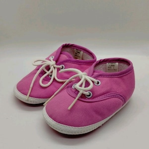 Könnte beinhalten: Ein Paar rosa Babyschuhe mit weißen Schnürsenkeln und weißer Sohle. Die Schuhe haben eine leinwandartige Textur und sind für Babys konzipiert. Größe 1.