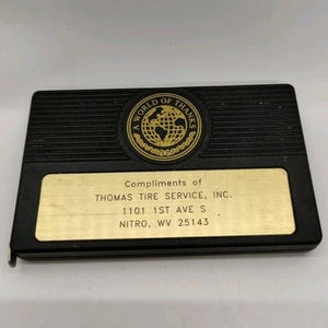 May include: Black rectangular tape measure with a gold-colored plate. The plate reads "Compliments of THOMAS TIRE SERVICE, INC." and includes an address. The top of the tape measure has a gold emblem with the words "A WORLD OF THANKS".