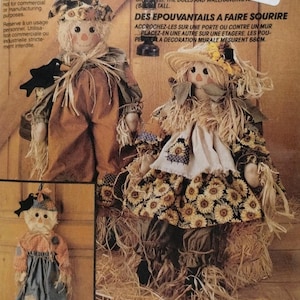 May include: Two fabric scarecrow dolls with straw hair and accents. One doll wears a brown outfit with a black bird, the other a sunflower print dress with a straw hat. Text includes Friendly Scarecrows and by/par Faye Wine. Dolls are 68 cm tall.