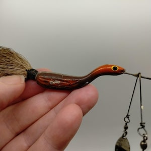 May include: A fishing lure shaped like a small bird with a brown and orange painted body, a yellow eye, and a tan feathered tail. The lure features a metal wire attachment with two dangling silver-coloured metal spinner blades for movement.
