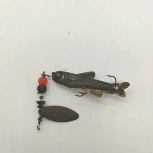 May include: A vintage fishing lure featuring a dark, textured fish-shaped body with two metal hooks, connected to a wire leader with a bright orange bead, a dark brown bead, and an oval-shaped metallic spinner blade with visible text markings.