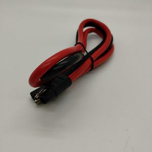 Puede incluir: Cable eléctrico rojo y negro con un conector negro. El cable está enrollado y asegurado con una brida negra. La imagen está tomada sobre un fondo blanco.