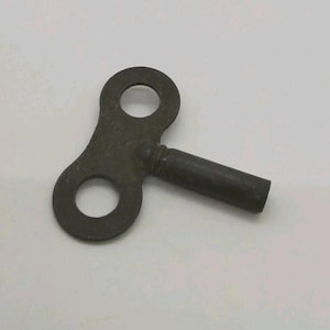 May include: A dark, weathered metal clock winding key featuring a flat, figure-eight shaped handle with two circular cutouts and a cylindrical shaft extending from the center, showing signs of oxidation and age on its surface.