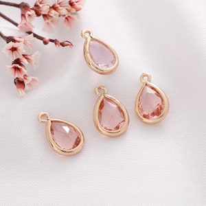 May include: Four gold-tone teardrop-shaped pendants with pink glass inserts. The pendants are on a white surface.