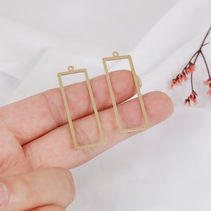 May include: Two gold-colored metal rectangular frames with a loop at the top. The frames are empty and ready to be filled with resin or other materials.