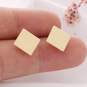 May include: A pair of gold-toned stud earrings featuring a square shape with a brushed, matte finish. The earrings have a flat, geometric design with clean edges and a metallic texture, presenting a simple and uniform appearance for each piece.