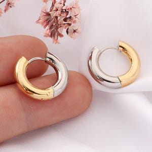 May include: Two-tone hoop earrings. Each earring features a gold-colored half and a silver-colored half. The earrings are round and have a hinged closure. The background is a soft white fabric with a few pink and brown flowers.