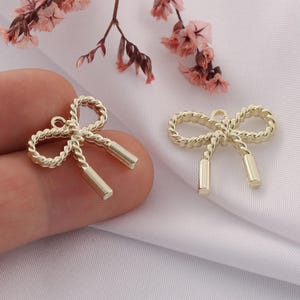 May include: Two gold-toned metal bow charms with a rope-like design. The bows have a loop at the top for attaching to a necklace, bracelet, or other jewelry.