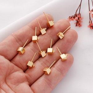 May include: Ten gold-toned cube earring posts with a loop on the side. The posts are on a hand.