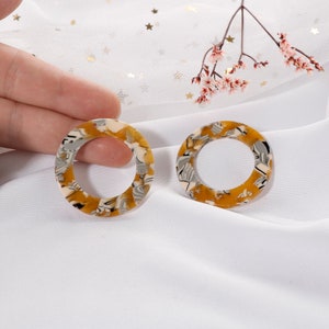 May include: A pair of large, round, yellow, white, and black patterned earrings. The earrings are made of resin and have a hole at the top for attaching to a hook.