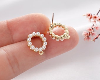 May include: A pair of gold-colored stud earrings. Each earring features a circular design, adorned with small, white pearls. The earrings are held by a person's fingers, showcasing the delicate craftsmanship and elegant style.