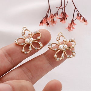May include: Two gold-tone flower-shaped charms with a pearl center and clear rhinestones. The charms have a loop at the top for attaching to jewelry or other crafts.