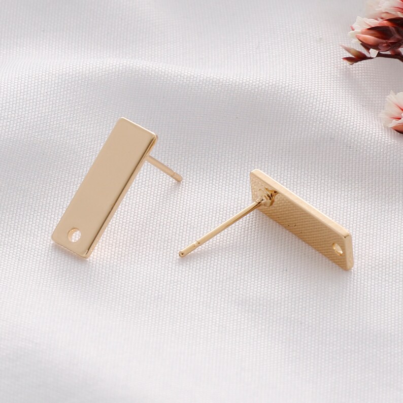 K&ouml;nnte beinhalten: Zwei goldfarbene rechteckige Ohrstecker. Jeder Ohrring hat einen Stift zum Einsetzen und ein kleines Loch unten. Die Ohrringe werden auf einer wei&szlig;en, strukturierten Oberfl&auml;che mit getrockneten Blumen im Hintergrund pr&auml;sentiert.