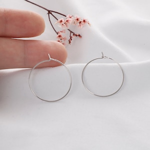May include: Two silver-colored hoop earrings. The earrings are circular and have a simple, minimalist design. The hoops are thin and appear to be made of metal. The background is a white, textured fabric, and a hand is holding one of the earrings.