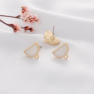 May include: A pair of gold-tone earrings with white enamel half-circle shapes. The earrings have a loop on the bottom for attaching charms or other jewelry.