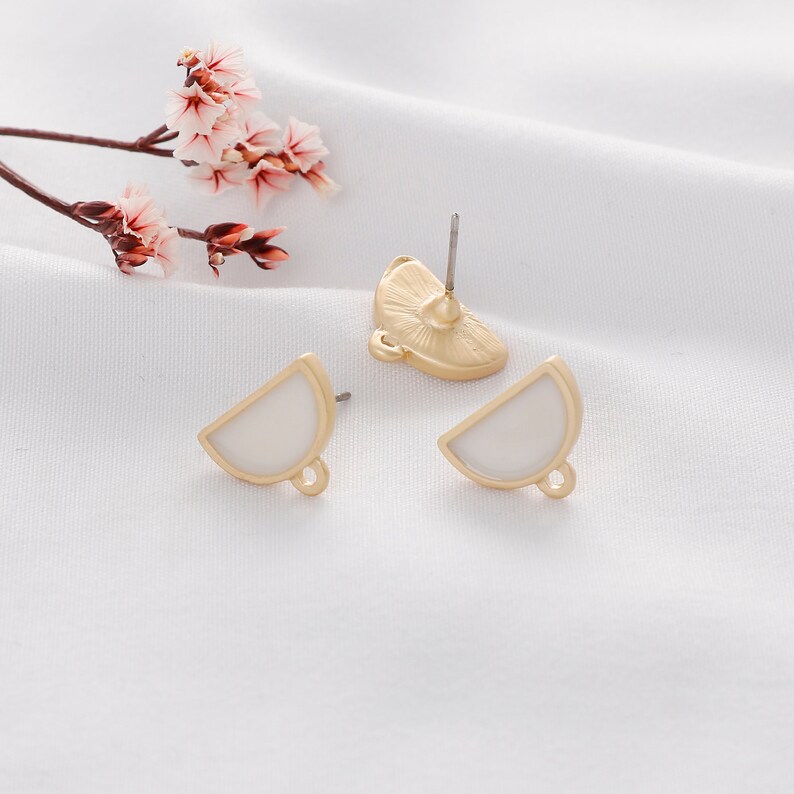 May include: A pair of gold-tone earrings with white enamel half-circle shapes. The earrings have a loop on the bottom for attaching charms or other jewelry.