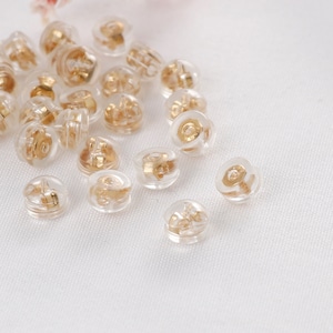 May include: A close-up of a group of clear plastic earring backs with gold-colored metal posts. The backs are scattered on a white surface.