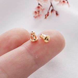 May include: Two gold-colored earring backs, also known as earring stoppers, are shown on a finger. The earring backs are small and round with a flat top.