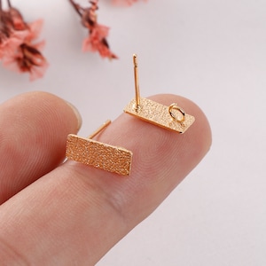 May include: Two gold-toned earring blanks with textured rectangular bases and a jump ring on one side. The earring blanks are on a finger.