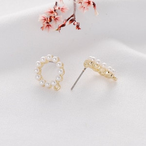 Puede incluir: Un par de pendientes de bot&oacute;n dorados, cada uno con un c&iacute;rculo de peque&ntilde;as perlas blancas. Los pendientes tienen un poste y un cierre. El fondo es una tela blanca suave.