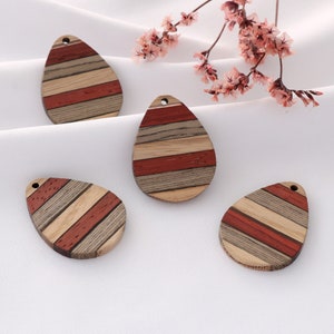 May include: Four teardrop-shaped wooden pendants with a hole at the top. Each pendant features a striped pattern of red, light brown, and gray wood. The pendants are arranged on a white surface, with pink flowers in the background.