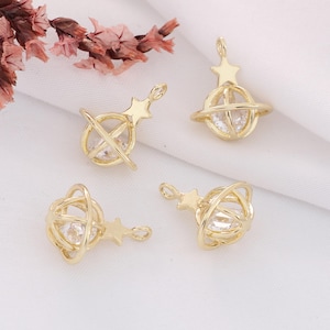 May include: Four gold-colored planet-shaped charms with a star and a clear crystal. The charms have a loop at the top for attaching to jewelry. The charms are on a white surface with a few pink flowers.