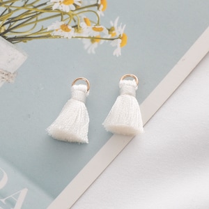 May include: Two white tassel earrings with gold-colored hoops. The tassels are made of soft, white fabric and are slightly flared at the bottom. The earrings are displayed on a light blue surface.