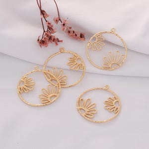May include: Several gold-colored, open-work circular pendants with a sunburst design. Each pendant has a small loop at the top for hanging. The pendants are displayed on a white, textured surface, with dried pink flowers in the background.